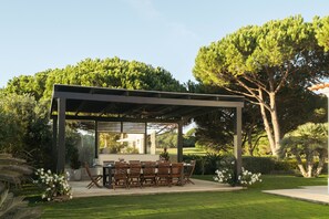 Eine Terrasse im Freien mit einer Pergola, einem Esstisch und Stühlen, umgeben von üppigem Grün und Bäumen.
