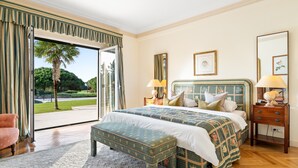 A spacious bedroom with a large bed, a bench, a nightstand, and a view of a garden through a sliding door. - Fairview Villa by Cadenzalux (Cascais)