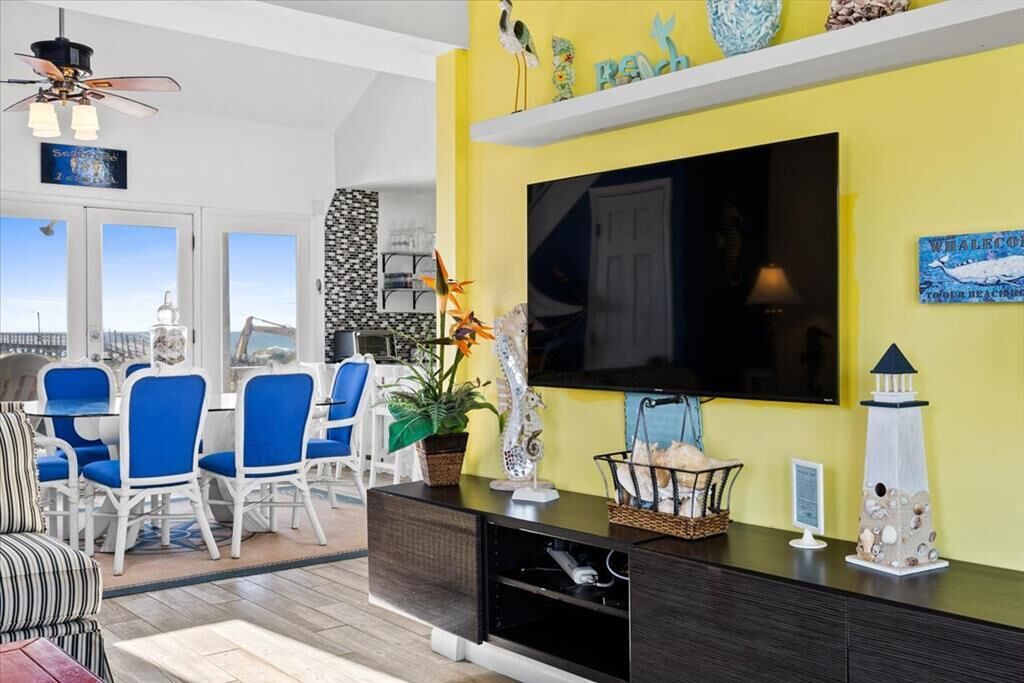 A bright yellow wall with a flat-screen TV, a dining area with blue chairs, and a ceiling fan.