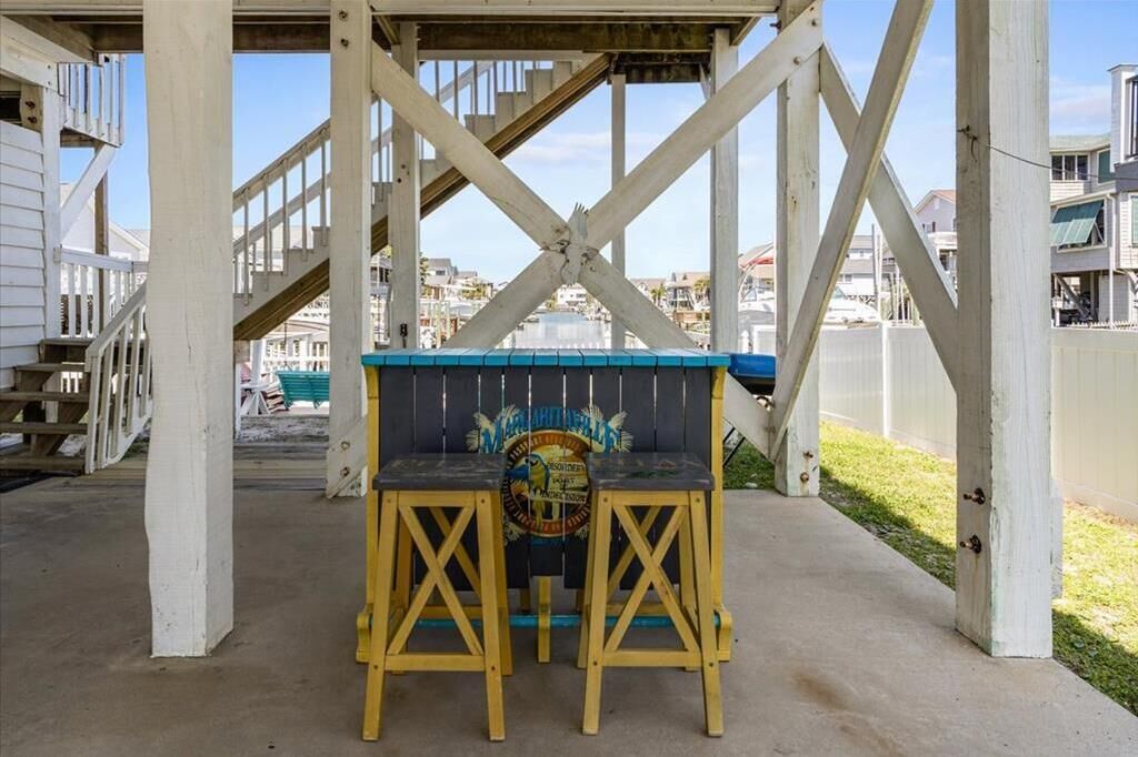 Un banco de madera bajo una estructura con techo azul, pilares blancos y vistas a casas al fondo.
