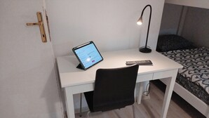 A small, white desk with a tablet and a remote control, a black chair, and a bed with patterned bedding.