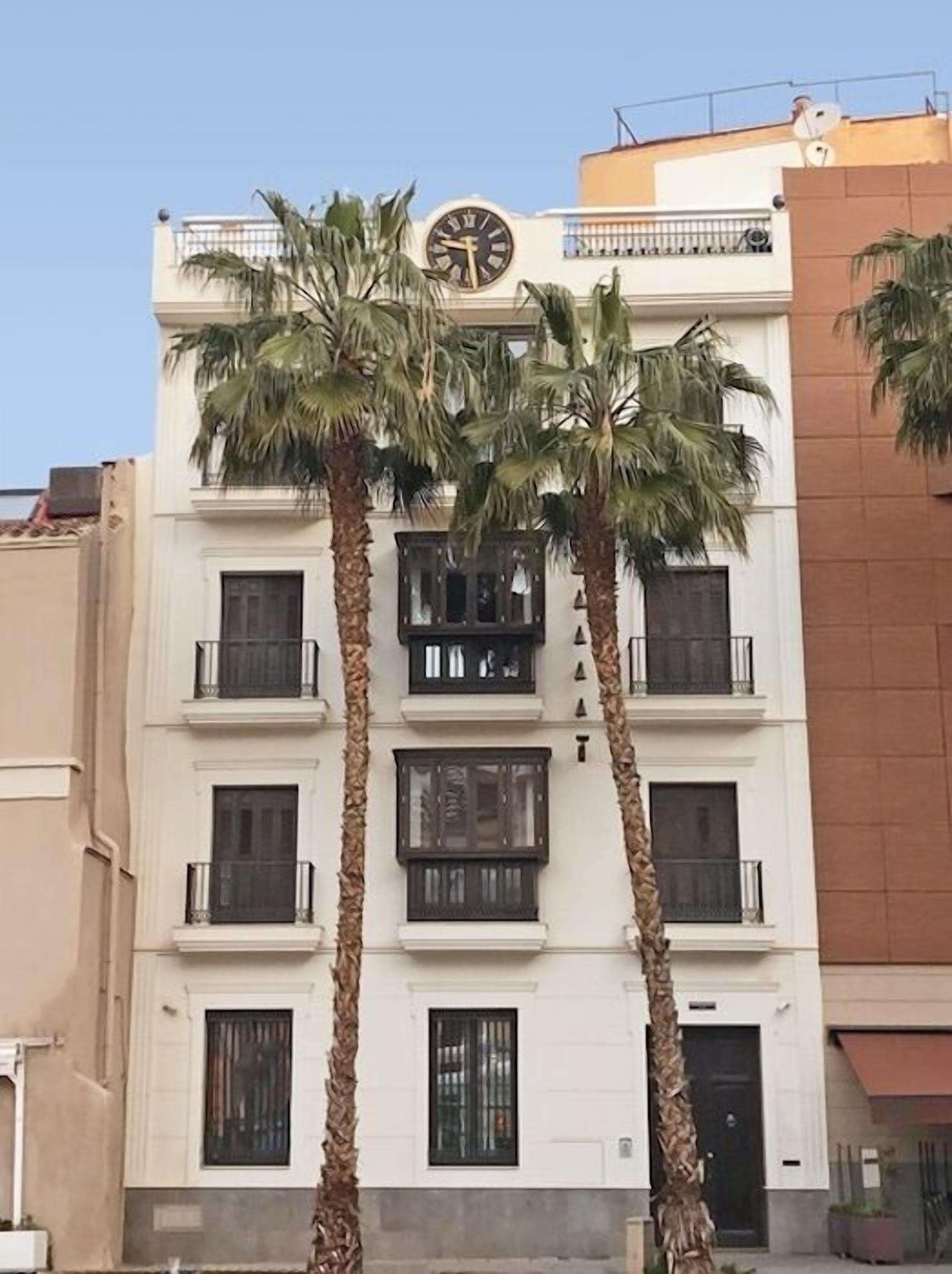 A multi-story building with a clock on the facade, balconies, and palm trees in front.