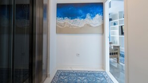 Un couloir orné d’un tableau abstrait aux couleurs bleue et blanche, une porte en verre et une étagère blanche sur laquelle sont disposés des objets décoratifs.
