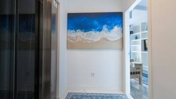 Un couloir orné d’un tableau abstrait aux couleurs bleue et blanche, une porte en verre et une étagère blanche sur laquelle sont disposés des objets décoratifs.