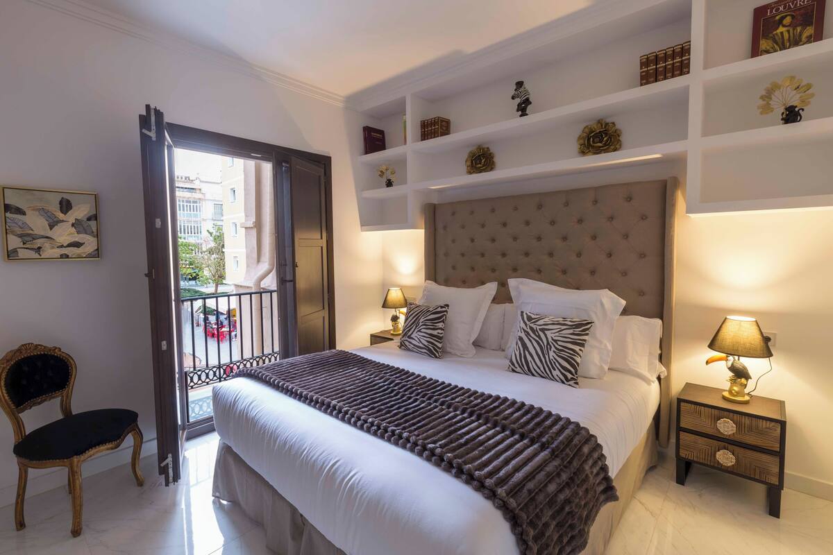 A bedroom with a large bed, a nightstand with a lamp, a chair, and a balcony with a view of buildings.