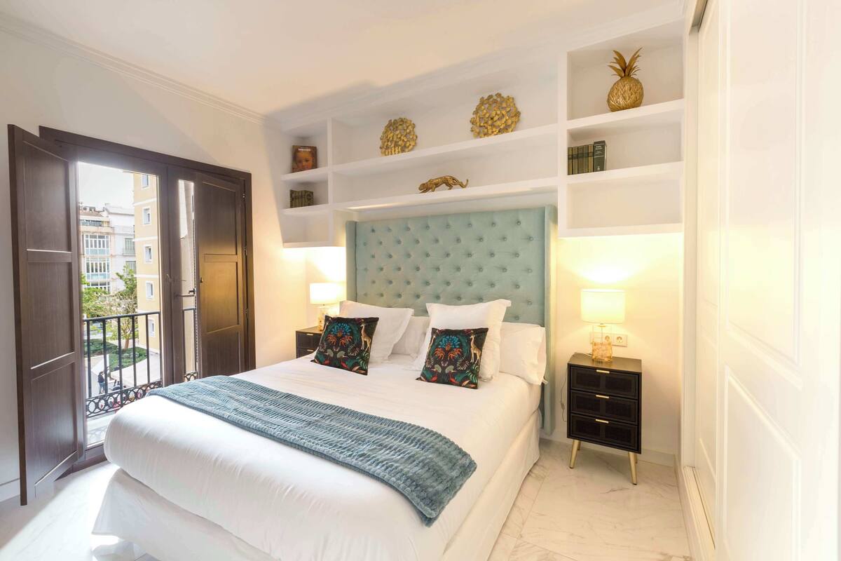 A hotel room with a large bed, a headboard with a tufted design, bedside tables with lamps, and a balcony with a view of buildings.