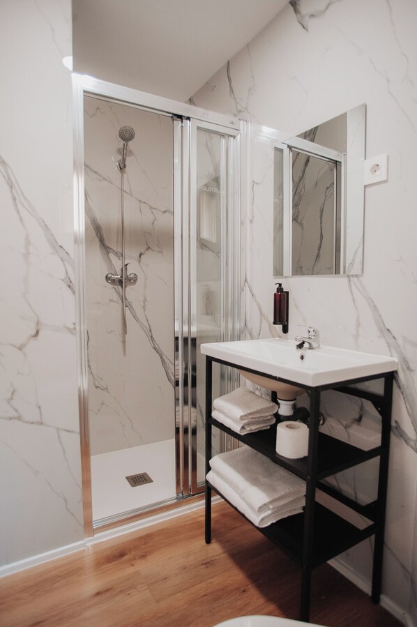 A bathroom with a glass-enclosed shower, a white sink, a mirror, and a shelf with towels. - Polotel (Valladolid)