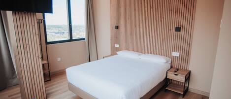 A modern bedroom with a large bed, a wooden slat wall, a window with a view, and a bedside table with a remote control.