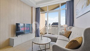 A modern living room with a large TV displaying "NETFLIX," a grey sofa, a small round coffee table, and a view of a balcony with a railing.