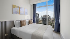 Een moderne hotelkamer met een groot bed, uitzicht over de stad en een raam met blauwe gordijnen.