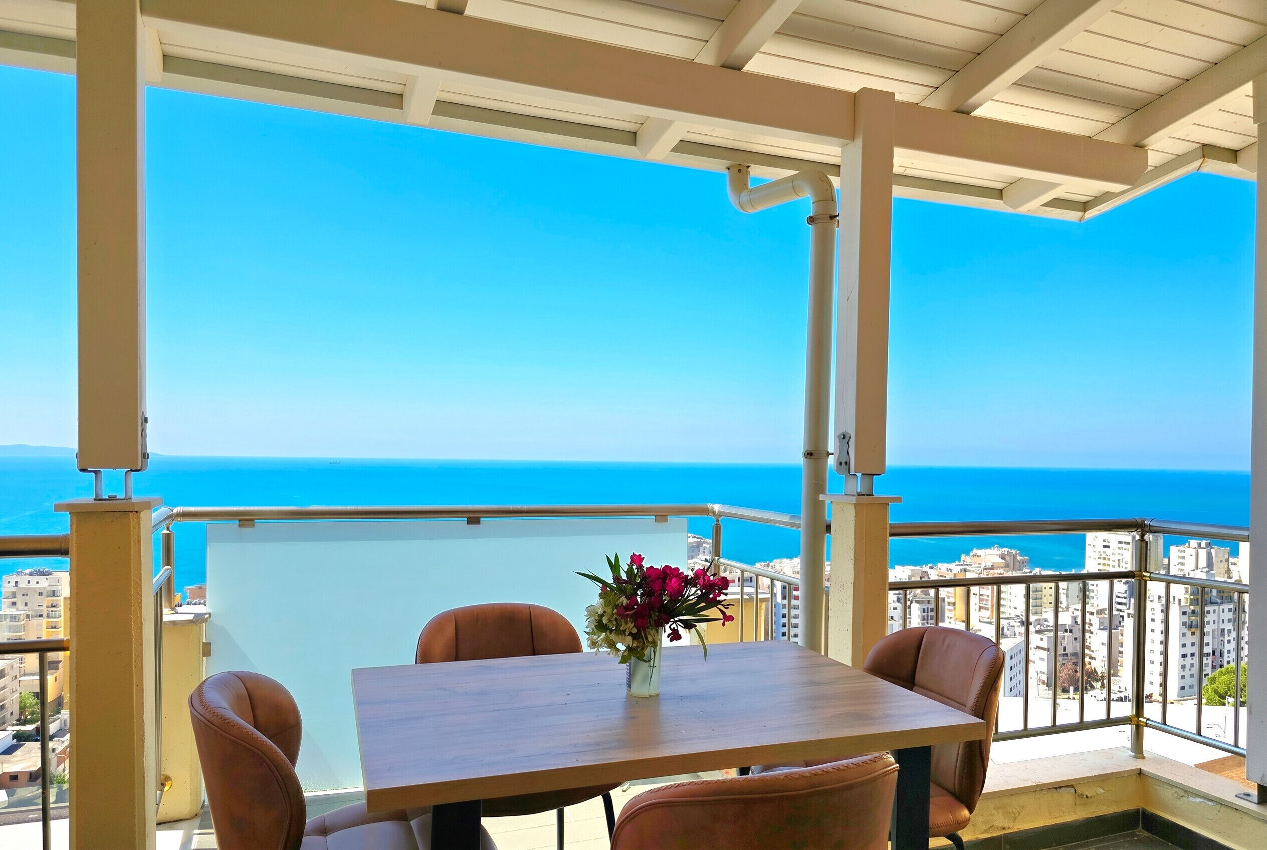 Un balcon avec une table et des chaises, un vase rempli de fleurs et une vue sur la mer et les bâtiments.
