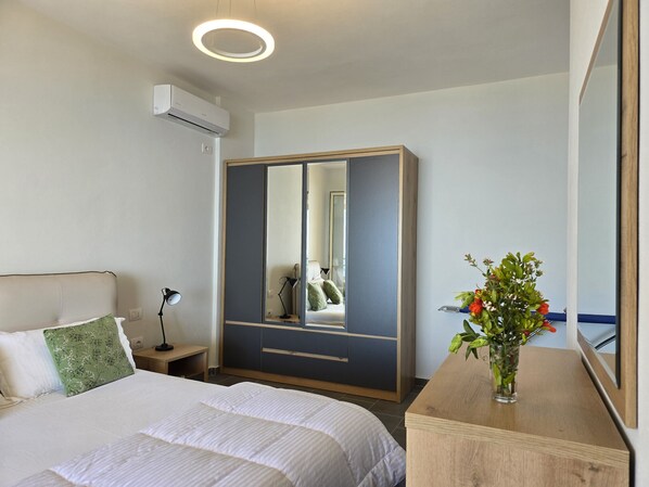 Quarto de hotel com cama, mesa de cabeceira, luminária, guarda-roupa, espelho e um vaso com flores.