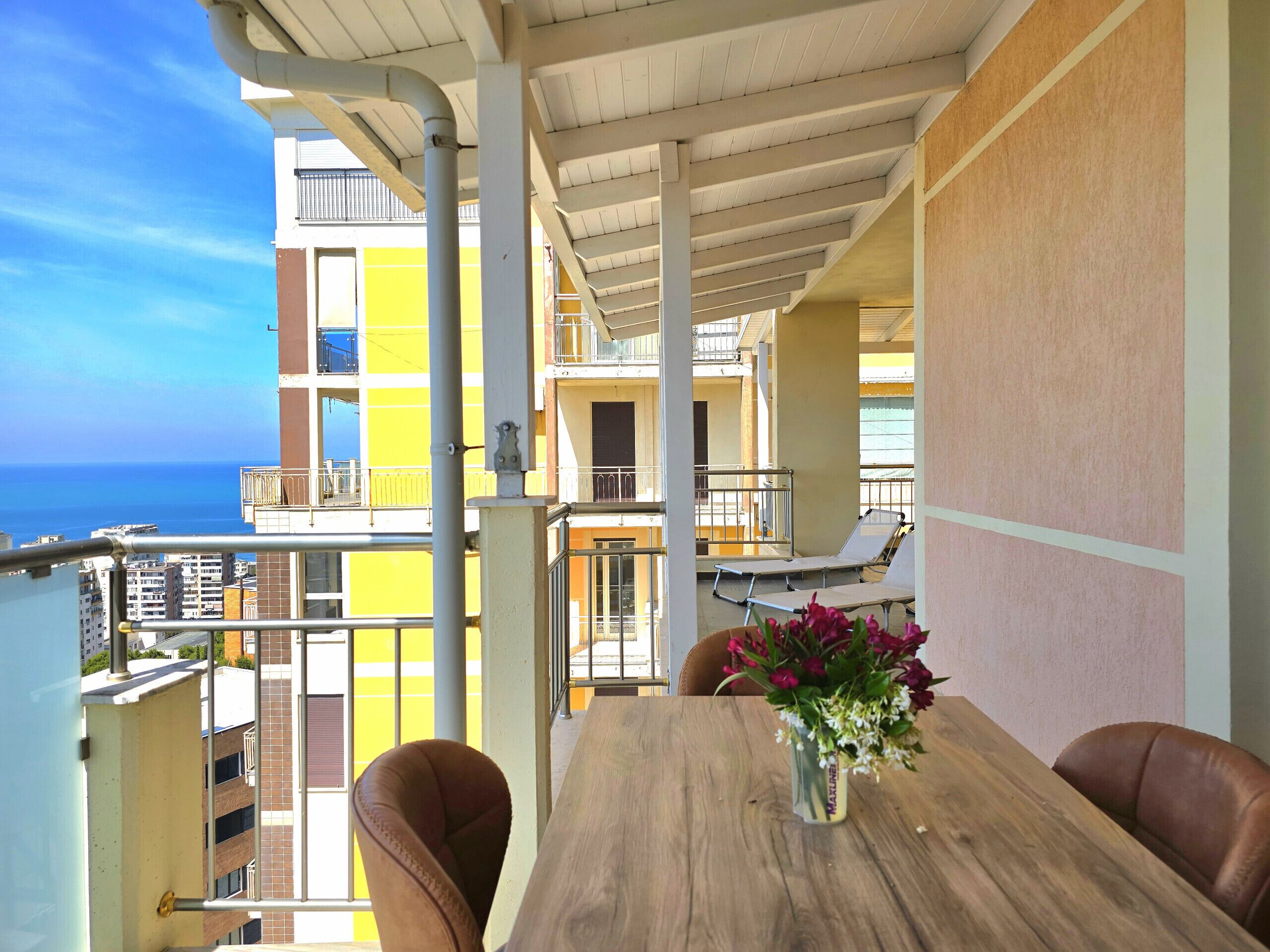 A balcony with a wooden table and chairs, a vase of flowers, and a view of the sea and buildings.
