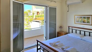 Une chambre à coucher comprenant un lit, une table de chevet, une lampe, un climatiseur, un ventilateur de plafond et une porte-fenêtre coulissante donnant sur un balcon.