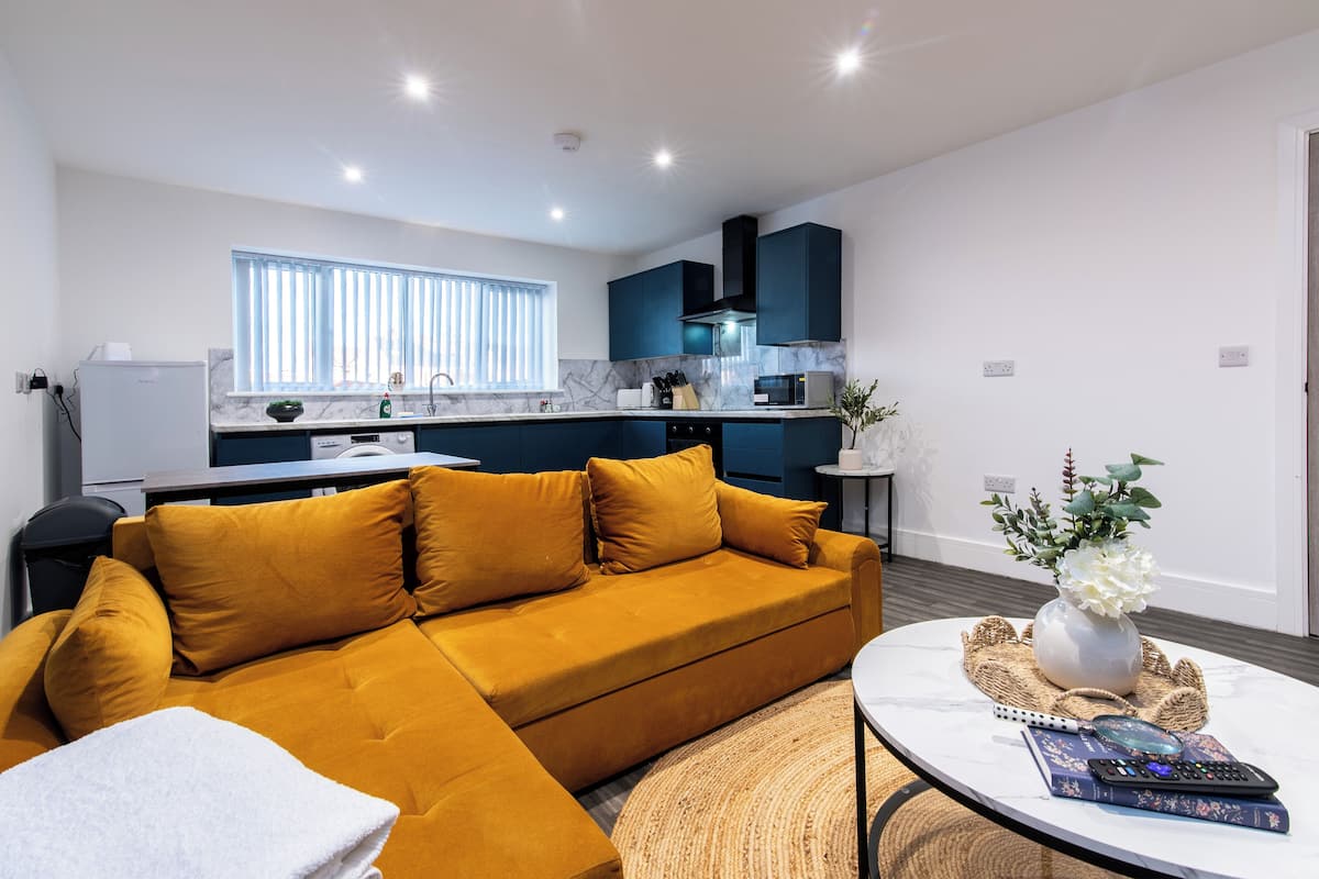 A modern living room with a mustard-colored sofa, a round coffee table with a vase and remote controls, and a kitchen in the background with blue cabinets and a window with blinds.