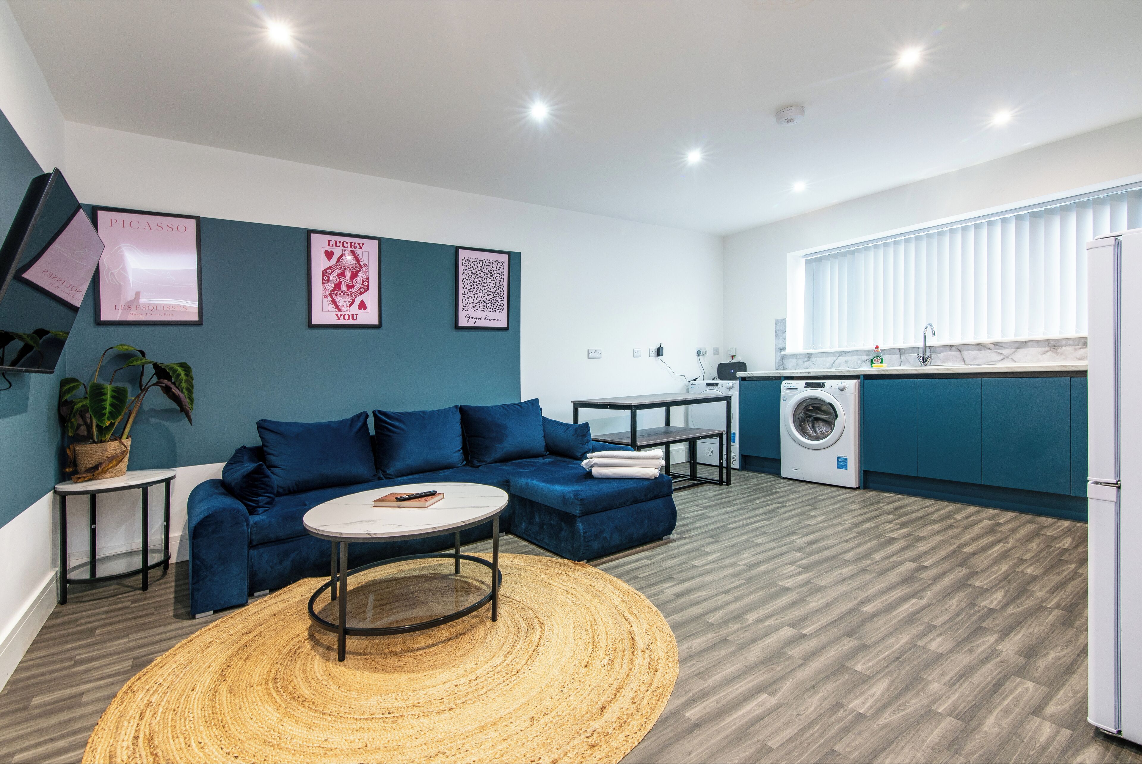 Un salon moderne avec un canapé bleu, une table basse ronde et un coin cuisine équipé d’un lave-linge.