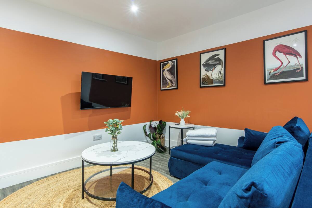 A modern living room with a large blue sofa, a small round table with a vase, and a flat-screen TV mounted on the wall.