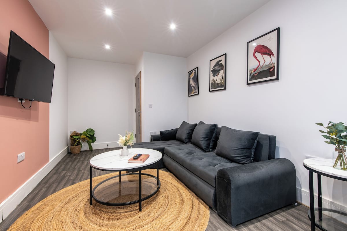 A modern living room with a black sofa, a round coffee table, and a flat-screen TV mounted on the wall. There are three framed artworks above the sofa and a potted plant on the floor.