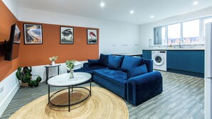 Un salon moderne avec un canapé bleu, une table basse ronde et un coin cuisine équipé d’un lave-linge.