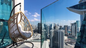 Une chaise en osier dotée d’un siège et d’un dossier rembourrés, placée sur un balcon dont la balustrade est en verre. Le balcon offre une vue sur un paysage urbain avec des immeubles modernes et des grues de construction.