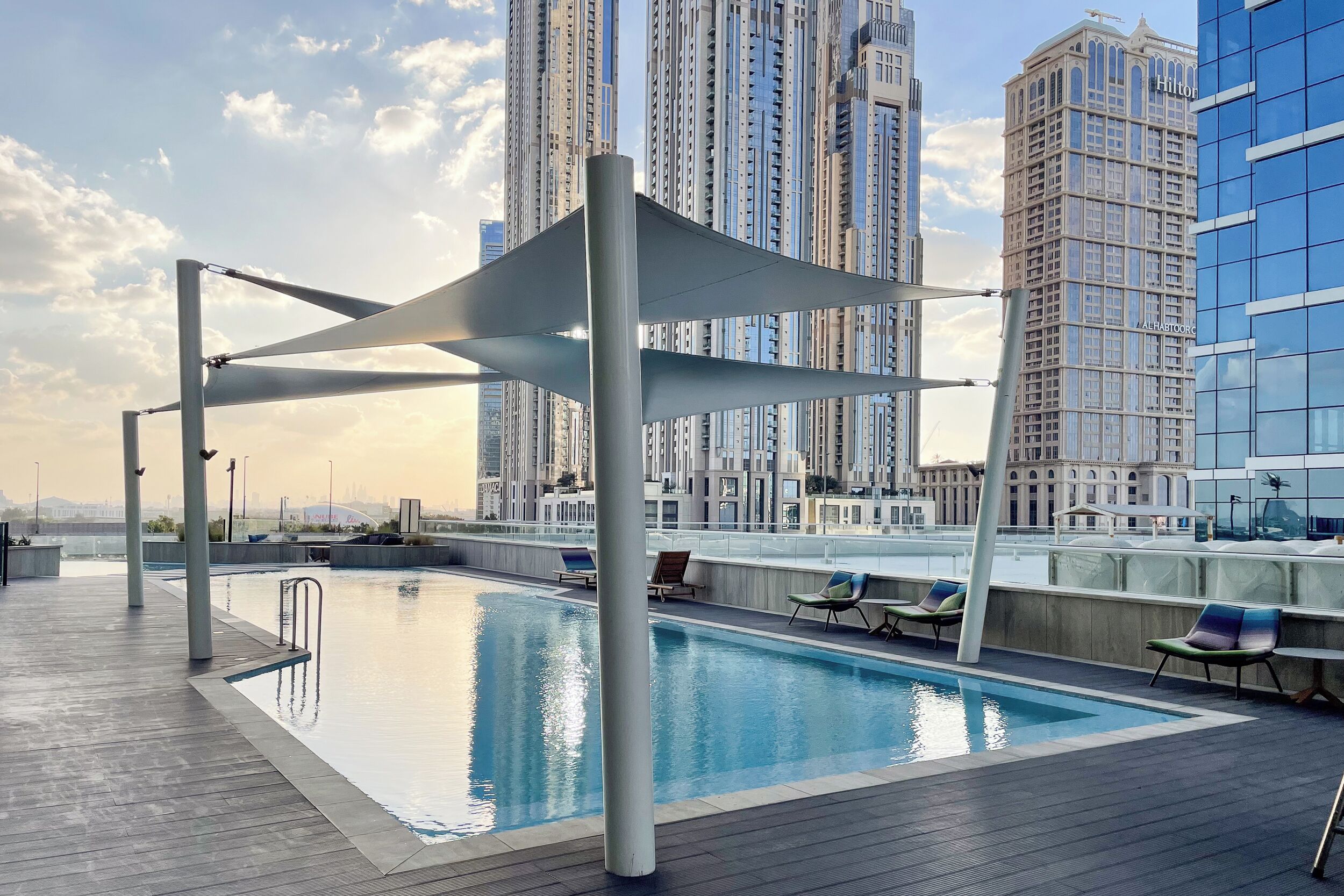 A rooftop swimming pool with lounge chairs and a canopy structure, surrounded by high-rise buildings.