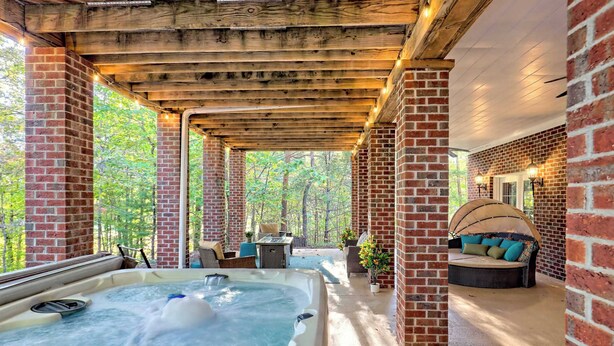 A hot tub in a covered outdoor area with brick walls and wooden beams, surrounded by seating and a view of trees. - Gold Leaf Chalet (Helen)