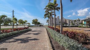 A paved pathway lined with palm trees and red flowering plants, leading to a residential area with modern buildings. - Vacation At Bac Bay! Fun In The Sun! 2B/2B Condo Walk To Beach! (Fort Myers Beach)