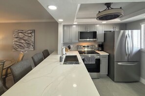A modern kitchen with a central island, stainless steel appliances, and a dining area with chairs. - Vacation At Bac Bay! Fun In The Sun! 2B/2B Condo Walk To Beach! (Fort Myers Beach)