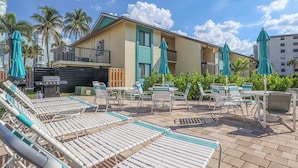 A poolside area with lounge chairs and umbrellas, a building with a balcony, and palm trees in the background. - Vacation At Bac Bay! Fun In The Sun! 2B/2B Condo Walk To Beach! (Fort Myers Beach)