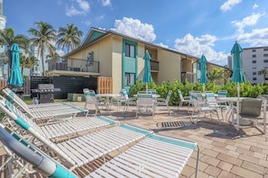 A poolside area with lounge chairs and umbrellas, a building with a balcony, and palm trees in the background. - Vacation At Bac Bay! Fun In The Sun! 2B/2B Condo Walk To Beach! (Fort Myers Beach)