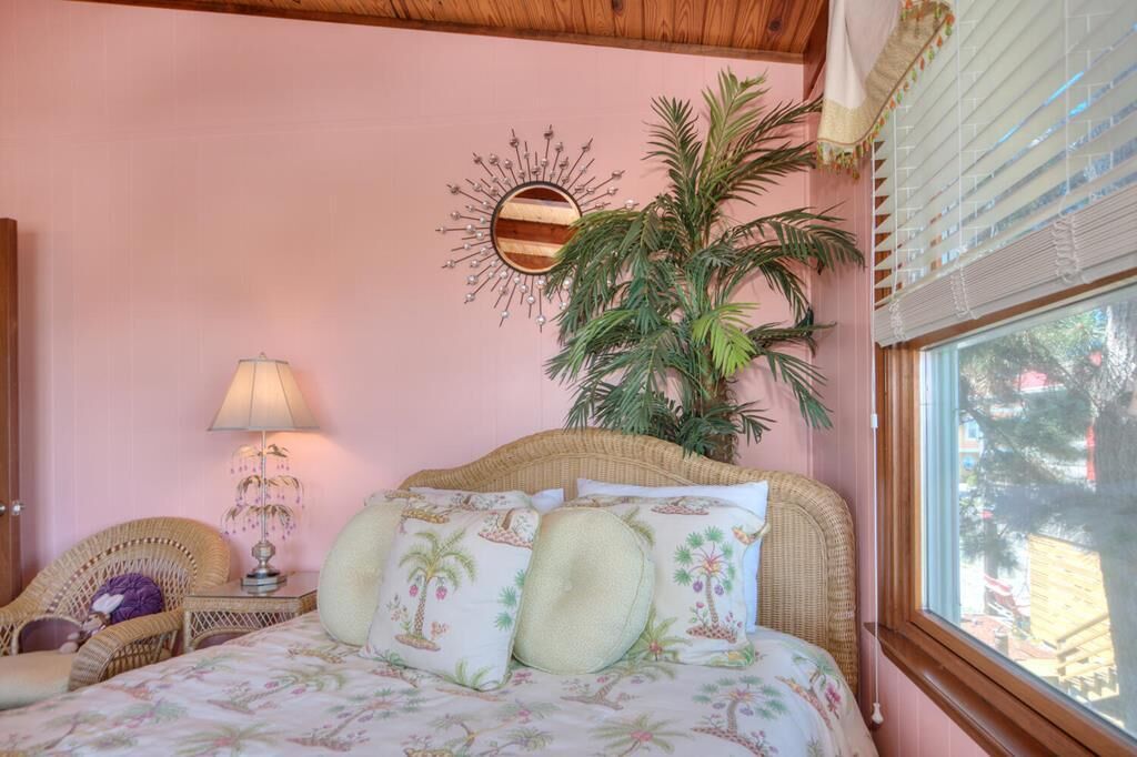 A bedroom with a bed, a wicker chair, a lamp, a decorative mirror, and a large plant.
