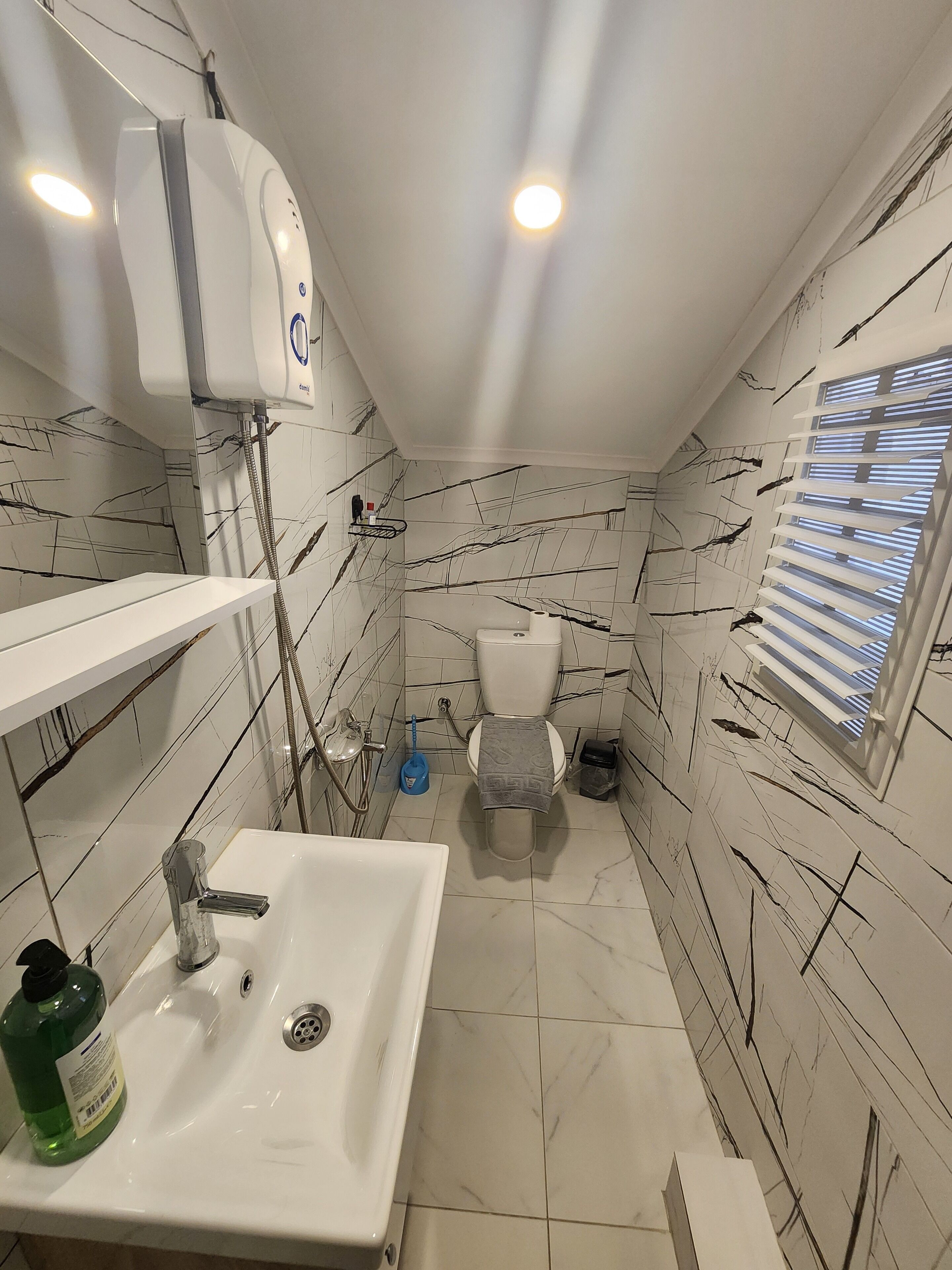 Ein Badezimmer mit einem weißen Waschbecken, einer Toilette und einem Duschbereich. Rechts befindet sich ein Fenster mit Jalousien.