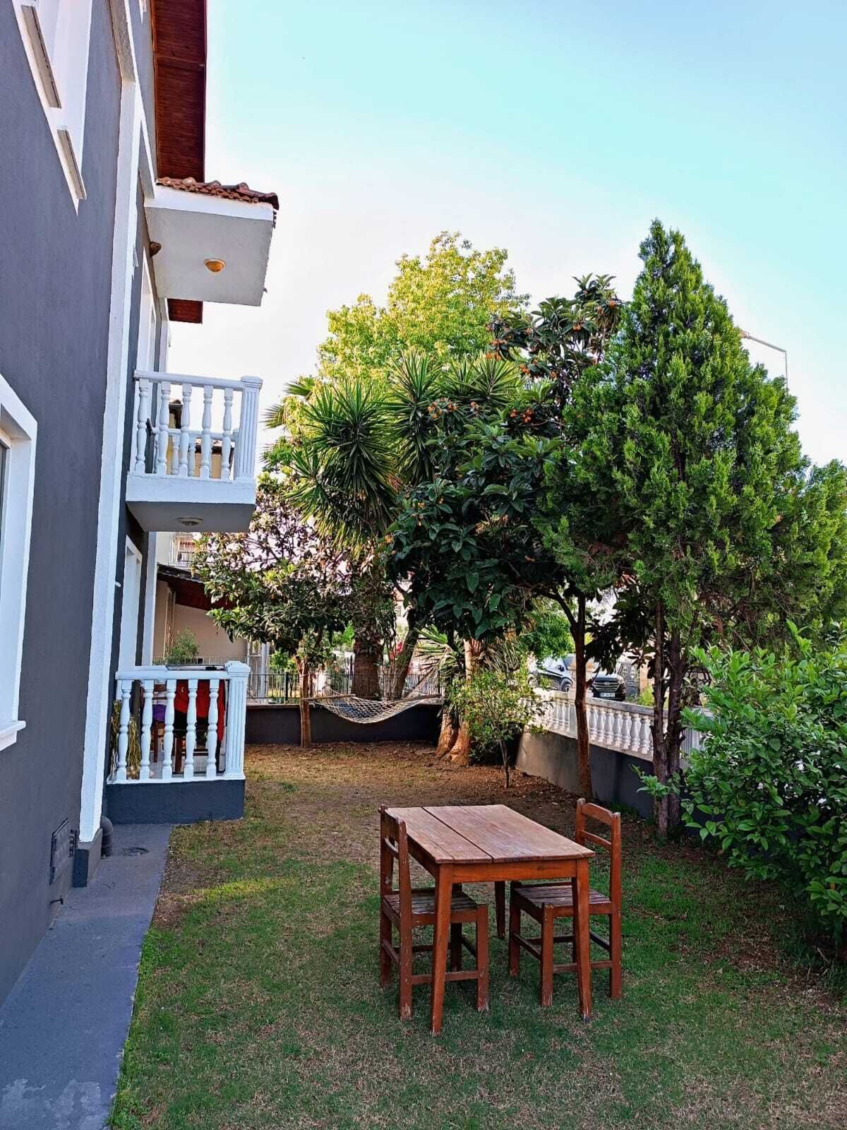 A small outdoor area with a wooden table and chairs, surrounded by greenery and a building with a balcony.
