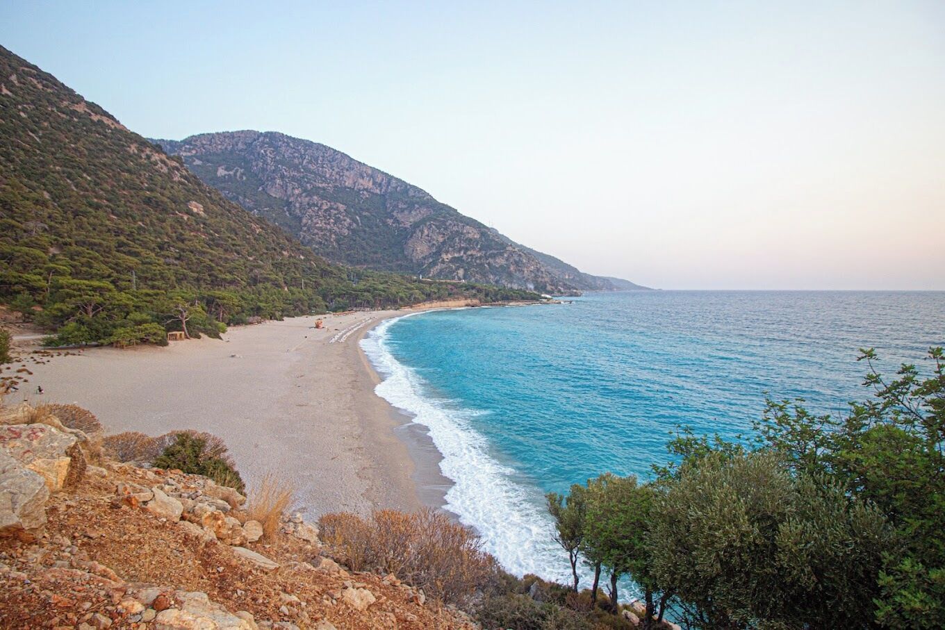 Paisatge costaner amb una platja, terreny rocos i una muntanya al fons.