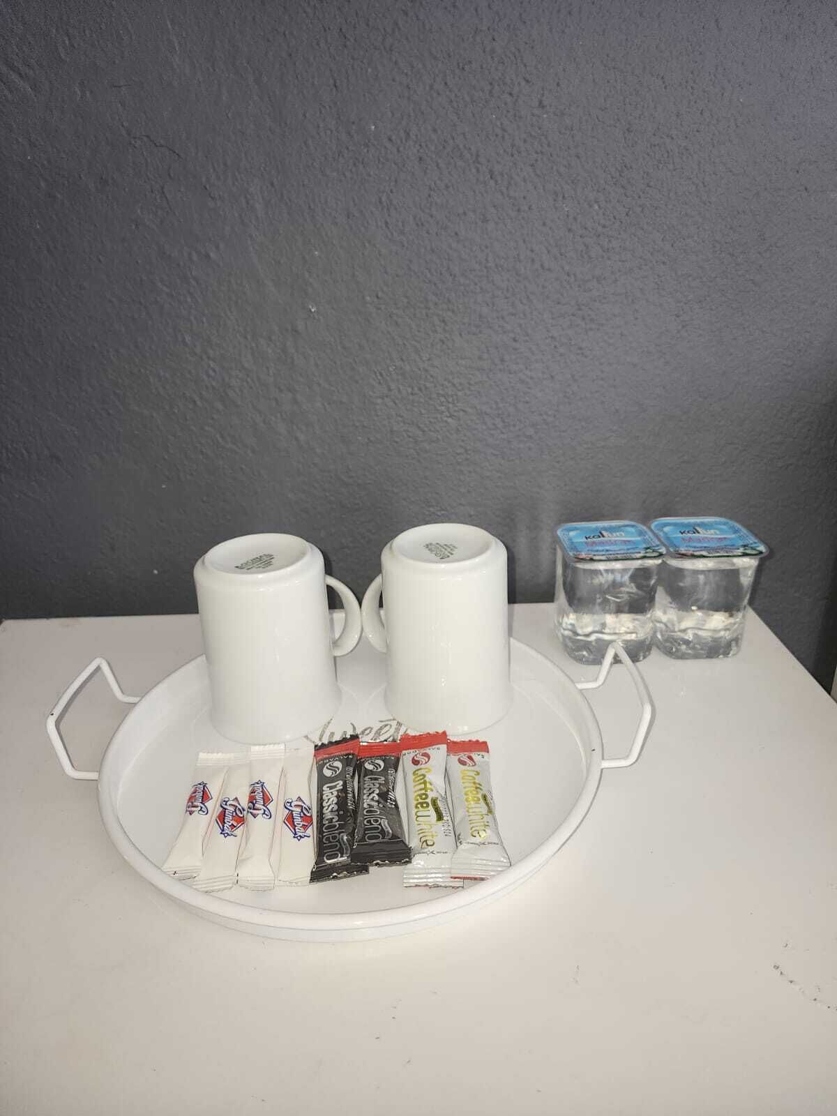 A tray with two white mugs, two glasses of water, and several packets of instant coffee on a white table against a grey wall.