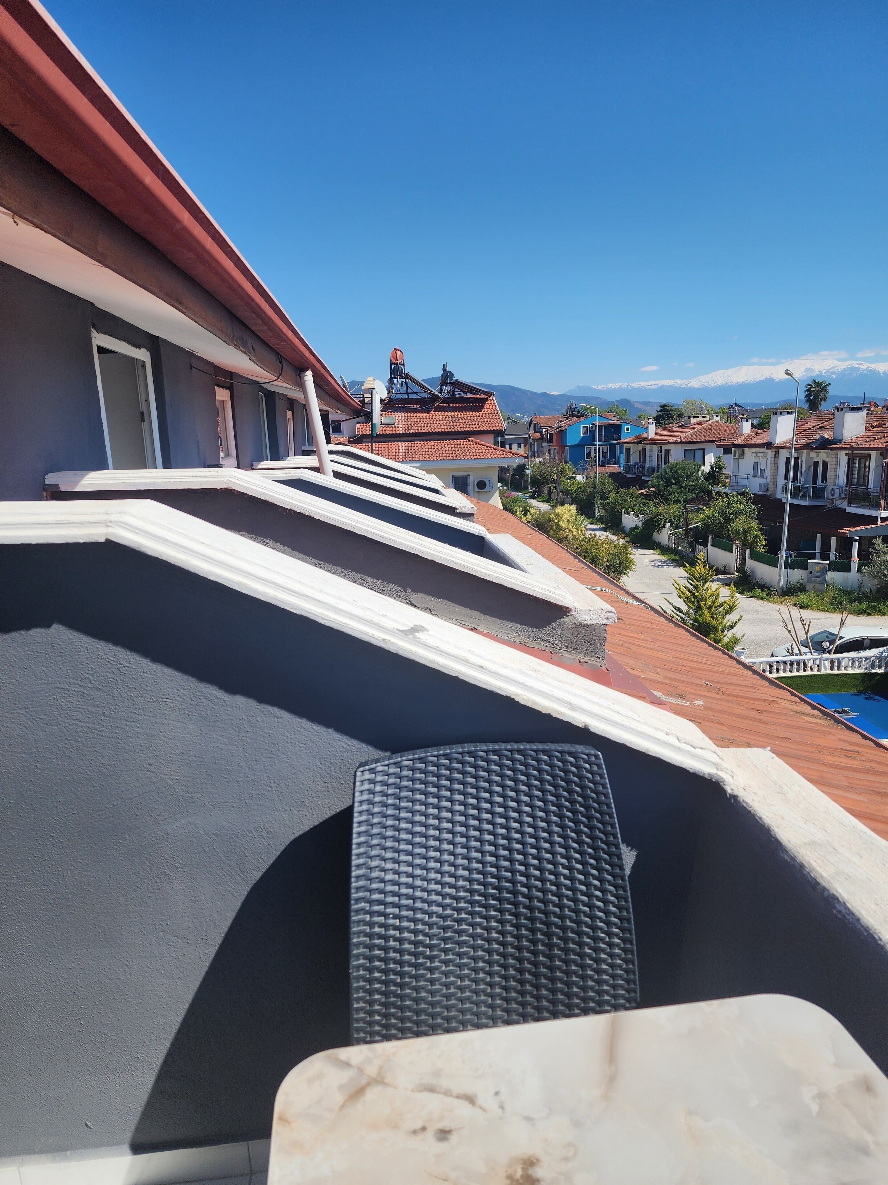 A rooftop terrace with a black textured floor, a white railing, and a view of neighboring buildings and mountains.