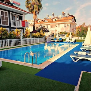 A swimming pool with lounge chairs, a white fence, and a building with a balcony in the background. - Şehzade Apart Otel (Fethiye)