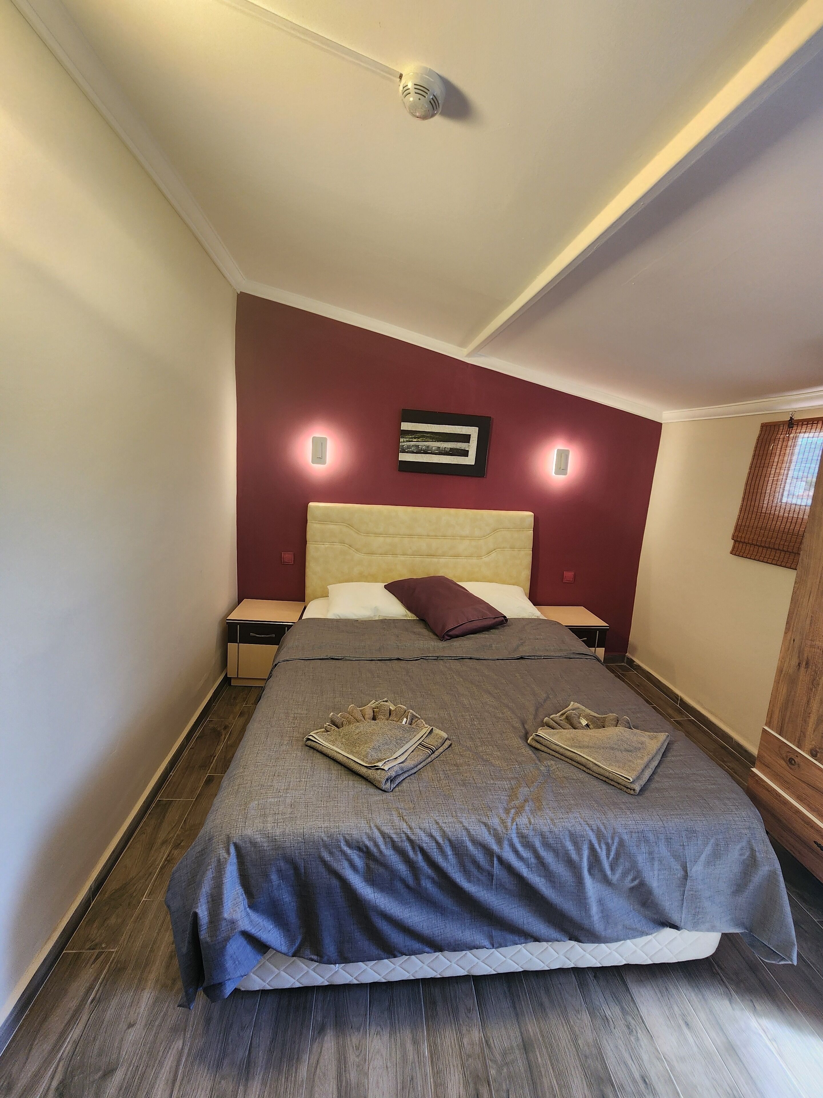 Cama bien tendida con una colcha gris, dos toallas dobladas y una sola almohada. Hay dos mesitas de noche, una con una lámpara y la otra con un enchufe. La habitación tiene una pared de acento en color rojo y una lámpara en el techo.