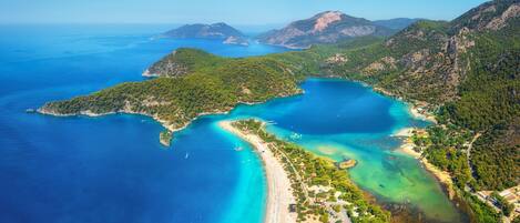 Vista aérea de una zona costera con una playa, aguas azules cristalinas y colinas verdes circundantes.