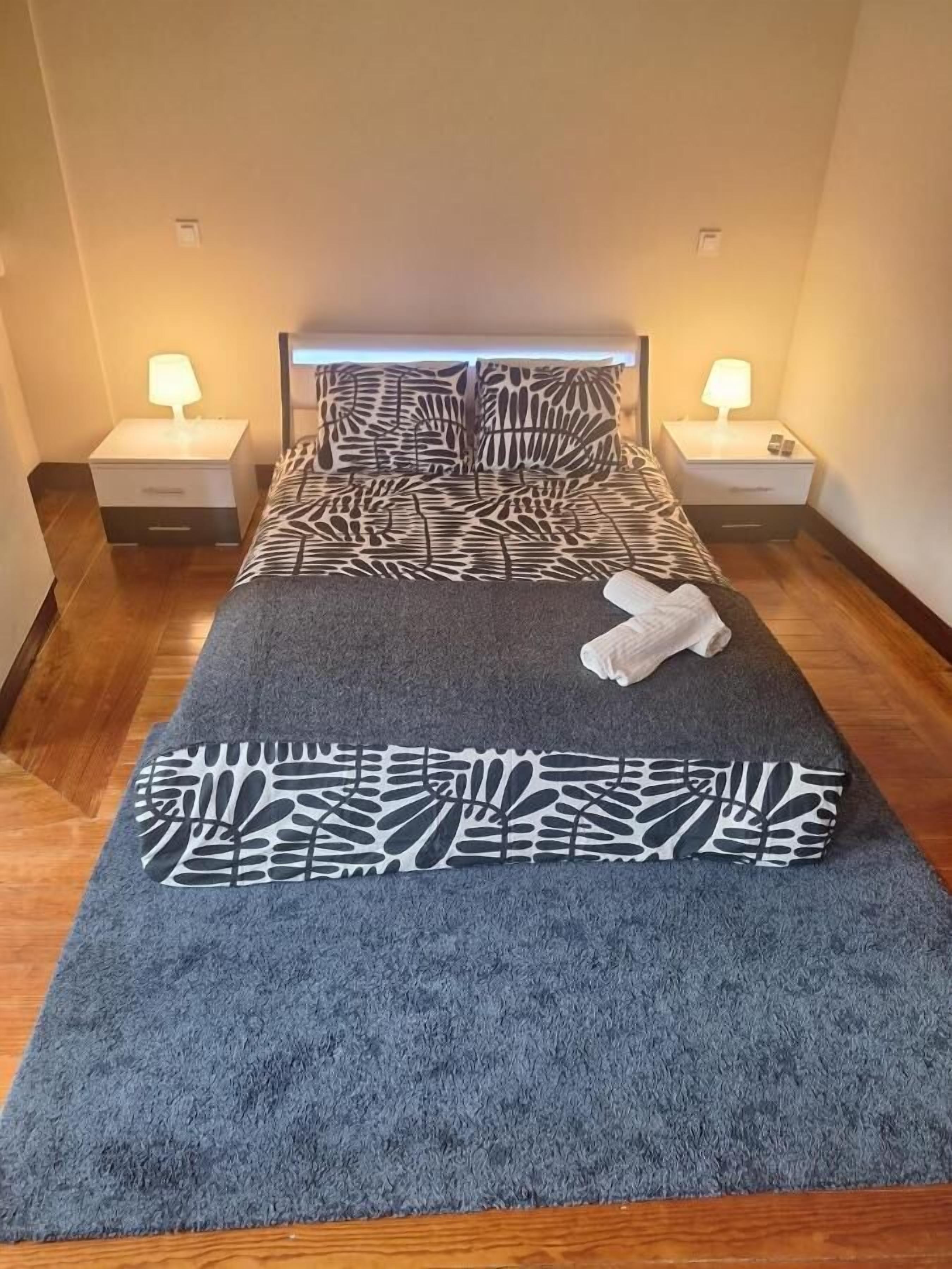Una cama bien tendida con una colcha estampada y almohadas, dos lámparas de noche y una toalla doblada sobre la cama.