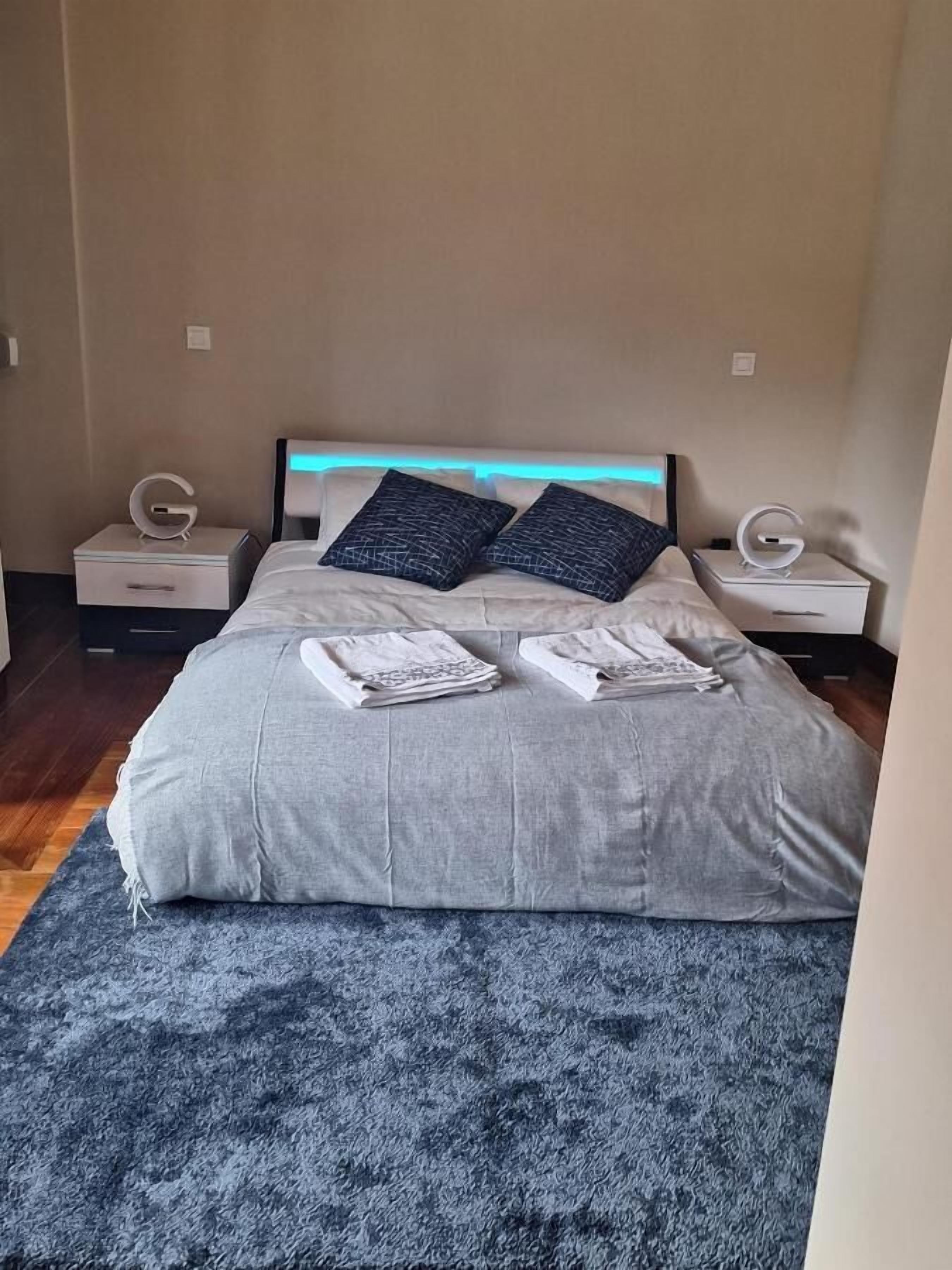 Una cama bien hecha con edredón gris y dos almohadas, una alfombra azul con textura y dos mesitas de noche con el logo blanco 'G'.