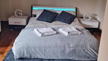 Una cama bien hecha con edredón gris y dos almohadas, una alfombra azul con textura y dos mesitas de noche con el logo blanco 'G'.