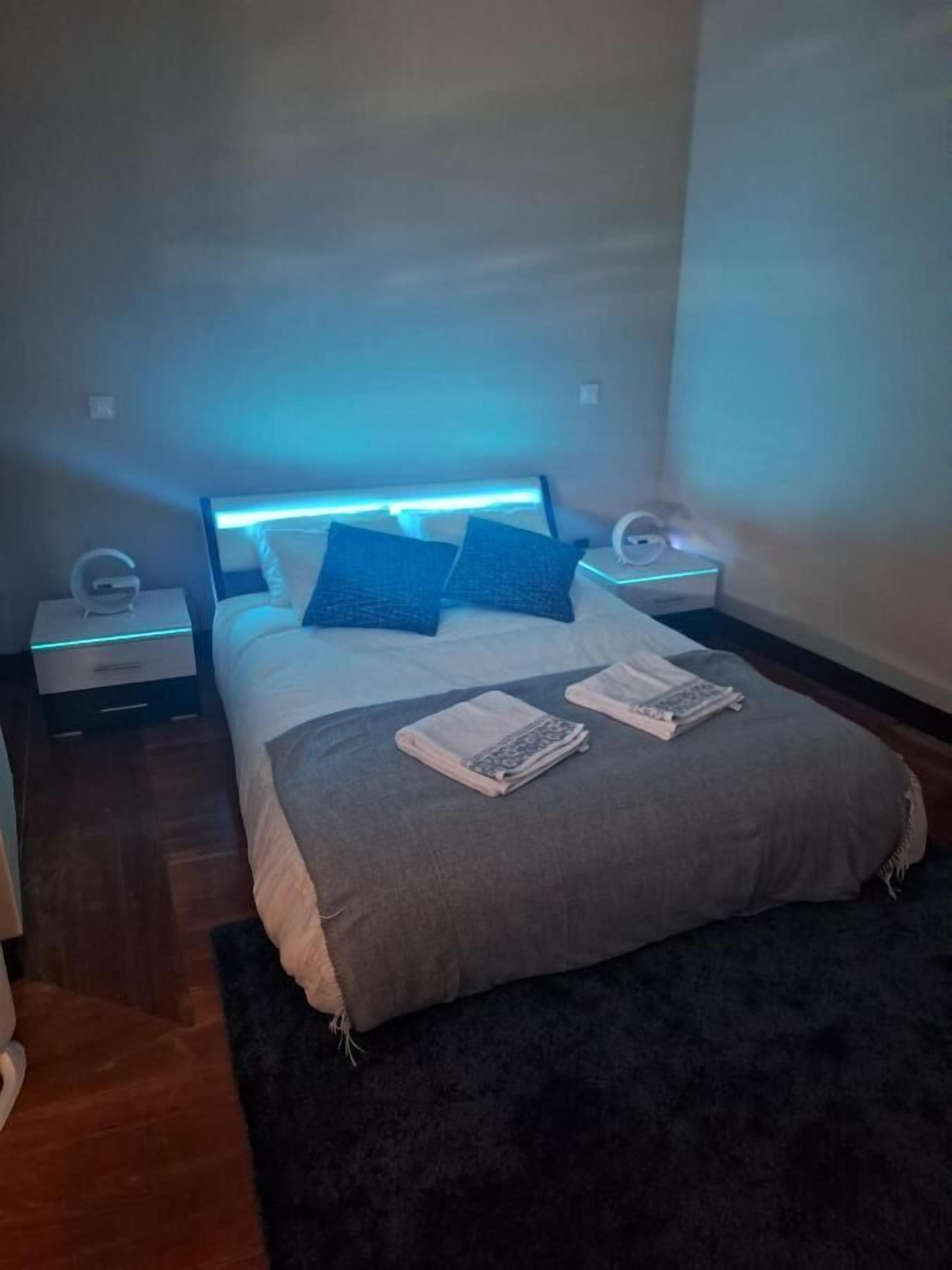 Un dormitorio con una cama, dos almohadas, una mesita de noche con una lámpara y un velador con un reloj.