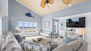 A spacious living room with a large window overlooking the beach, a ceiling fan, and a flat-screen TV. - Grand View, Ocean Isle Beach (Ocean Isle Beach)