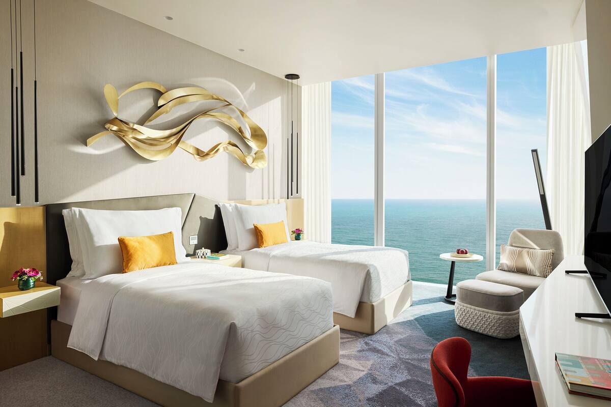 A modern hotel room with two beds, a desk, a chair, and a view of the ocean.