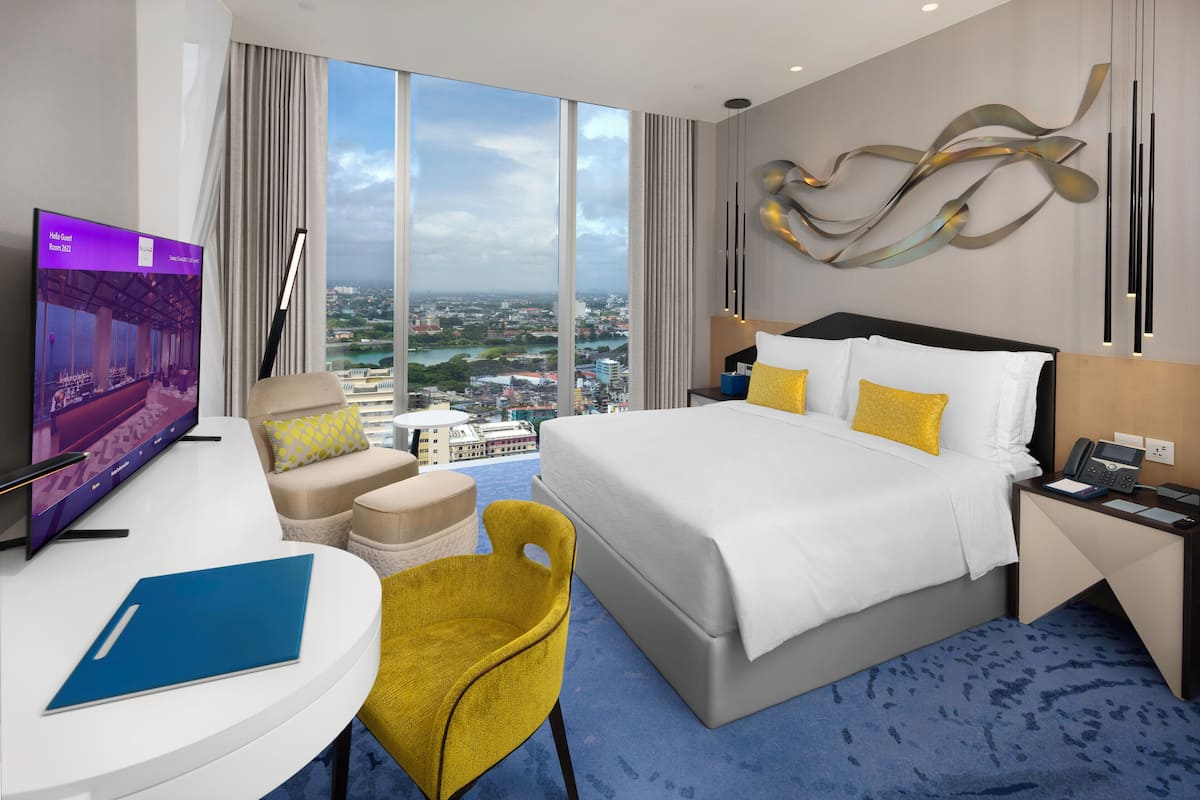 A hotel room with a large bed, a desk with a TV, a yellow chair, and a view of a cityscape.