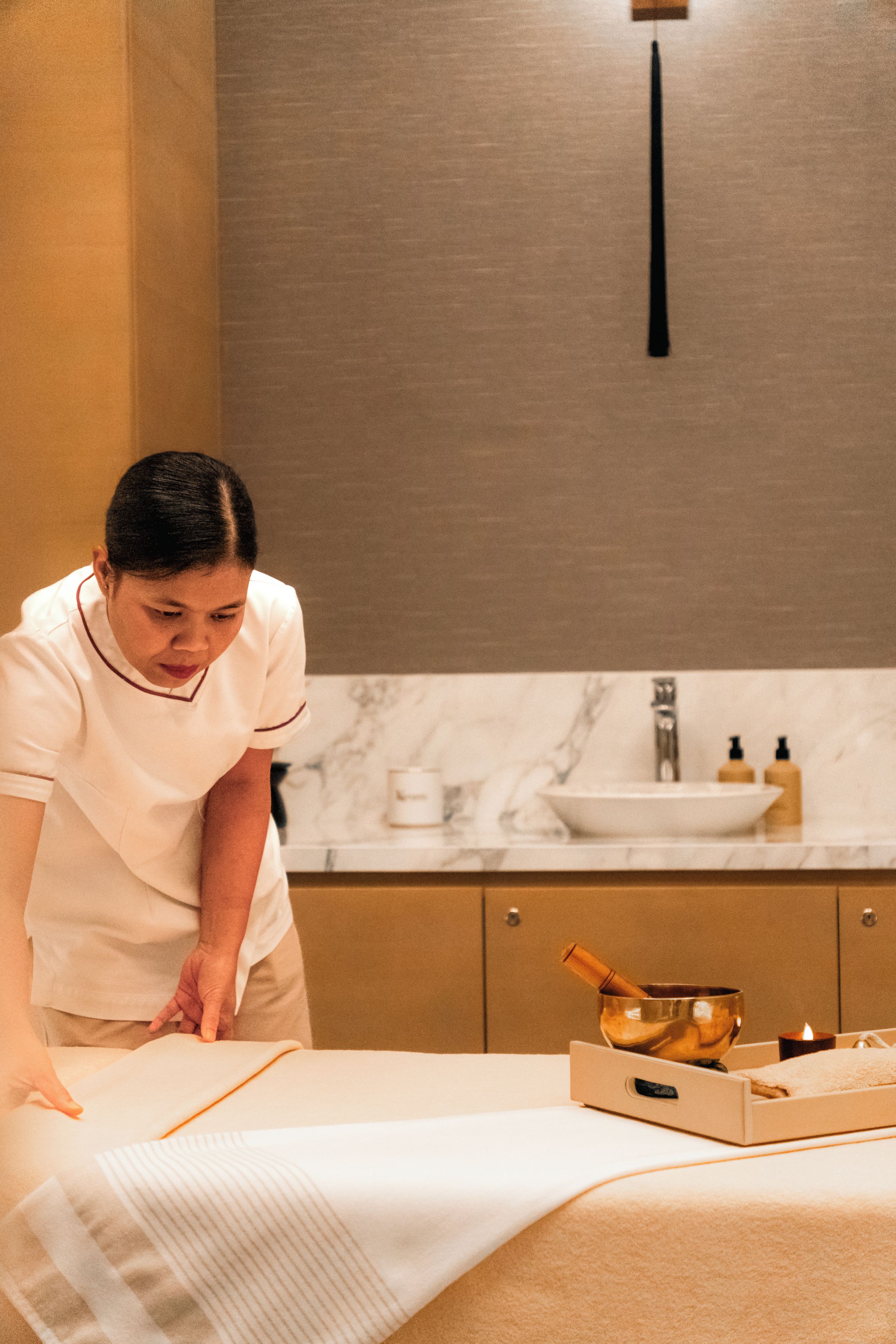 A person attending to a massage table with a towel and a bowl, in a room with a marble countertop and a wall-mounted light fixture.