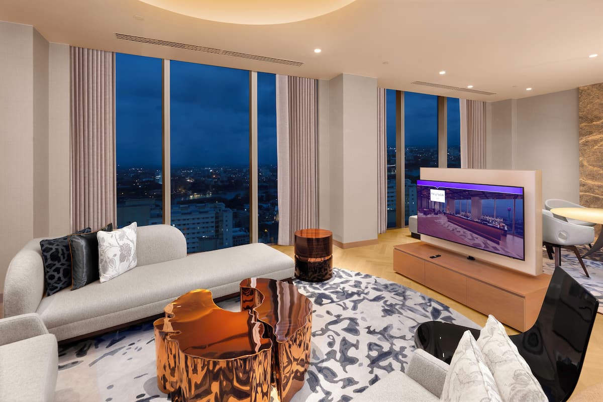 A modern living room with a large flat-screen TV, a sofa, a coffee table, and a view of a cityscape at dusk.