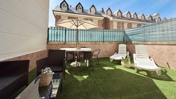 Eine Terrasse mit Tisch und Stühlen, ein kleiner Gartenbereich mit Kunstgrashalm und Blick auf ein Gebäude mit markanter Dachlinie.