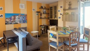 Ein gemütliches Wohnzimmer mit einer Couch, einem Esstisch mit Stühlen, einem Fernseher und einer Küchenzeile mit Schränken und einem Fenster.